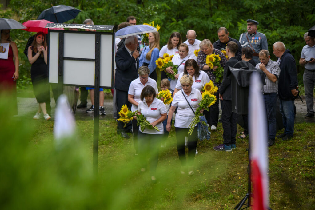 Grupa ludzi z kwiatami stoi na mokrej trawie pod parasolami. Ubrani w białe koszulki, w tle zielone drzewa. Tablica na pierwszym planie.