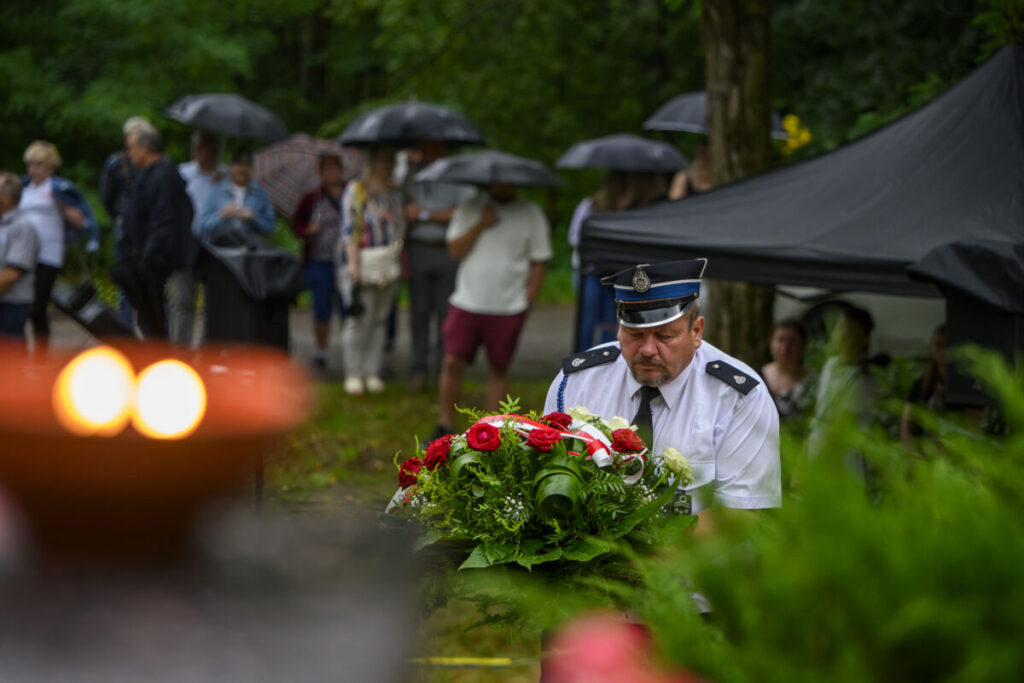 Mężczyzna w mundurze niesie wieniec z czerwonymi różami. W tle ludzie pod parasolami w deszczowej scenerii. Zieleń i namiot dominują w tle.