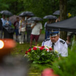 Mężczyzna w mundurze niesie wieniec z czerwonymi różami. W tle ludzie pod parasolami w deszczowej scenerii. Zieleń i namiot dominują w tle.