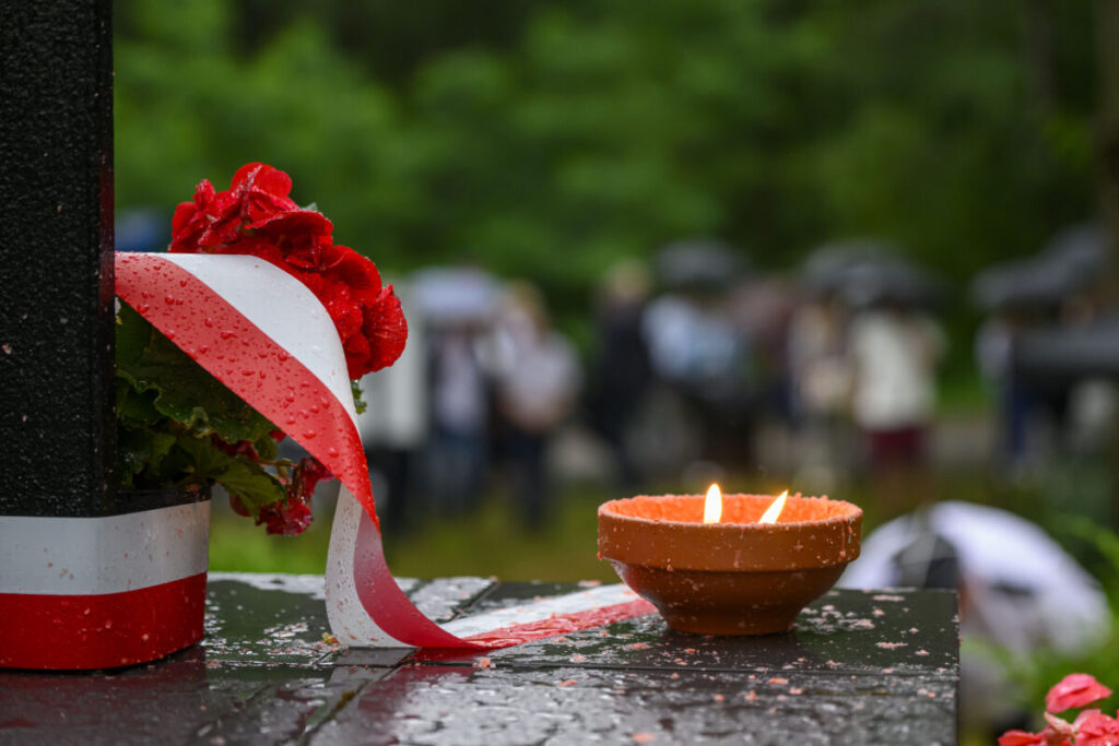 Bukiet czerwonych kwiatów z biało-czerwoną wstążką leży obok płonącego znicza. Tło zamglone, zielone drzewa i ludzie. Pada deszcz.