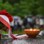 Bukiet czerwonych kwiatów z biało-czerwoną wstążką leży obok płonącego znicza. Tło zamglone, zielone drzewa i ludzie. Pada deszcz.