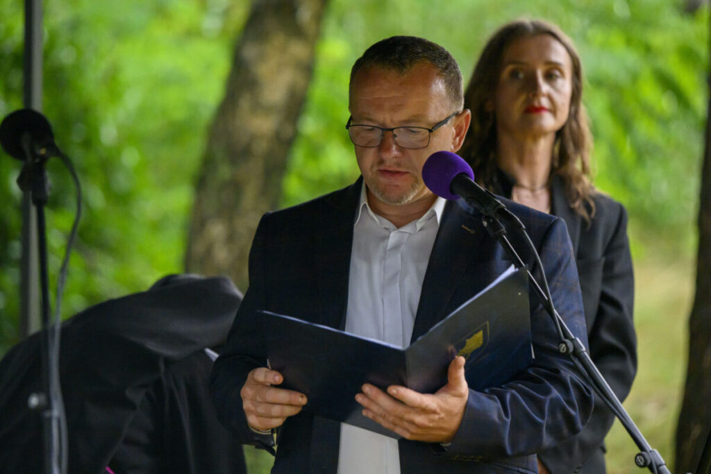 Mężczyzna w okularach czyta z kartki przy mikrofonie na tle zieleni. Obok stoi kobieta. Oboje ubrani są elegancko.