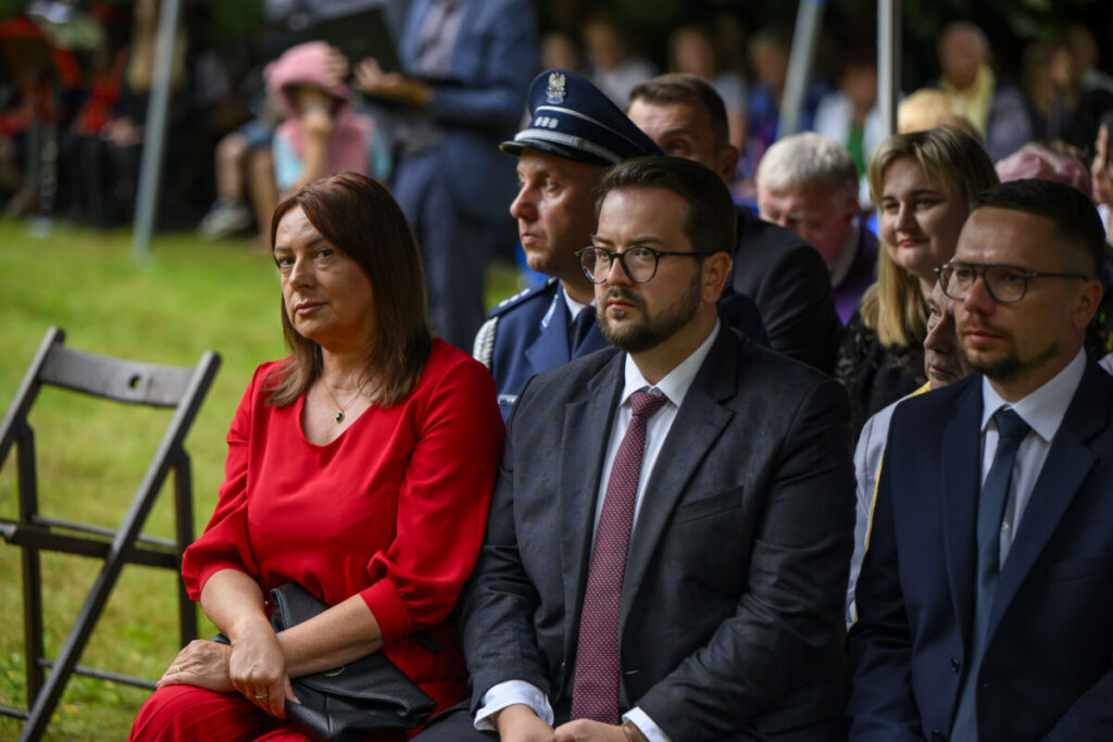 Grupa ludzi siedzi na zewnątrz w formalnym wydarzeniu. Kobieta w czerwonej sukience i mężczyźni w garniturach. Tło to trawnik i inni uczestnicy.