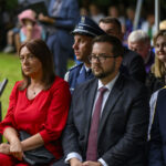 Grupa ludzi siedzi na zewnątrz w formalnym wydarzeniu. Kobieta w czerwonej sukience i mężczyźni w garniturach. Tło to trawnik i inni uczestnicy.