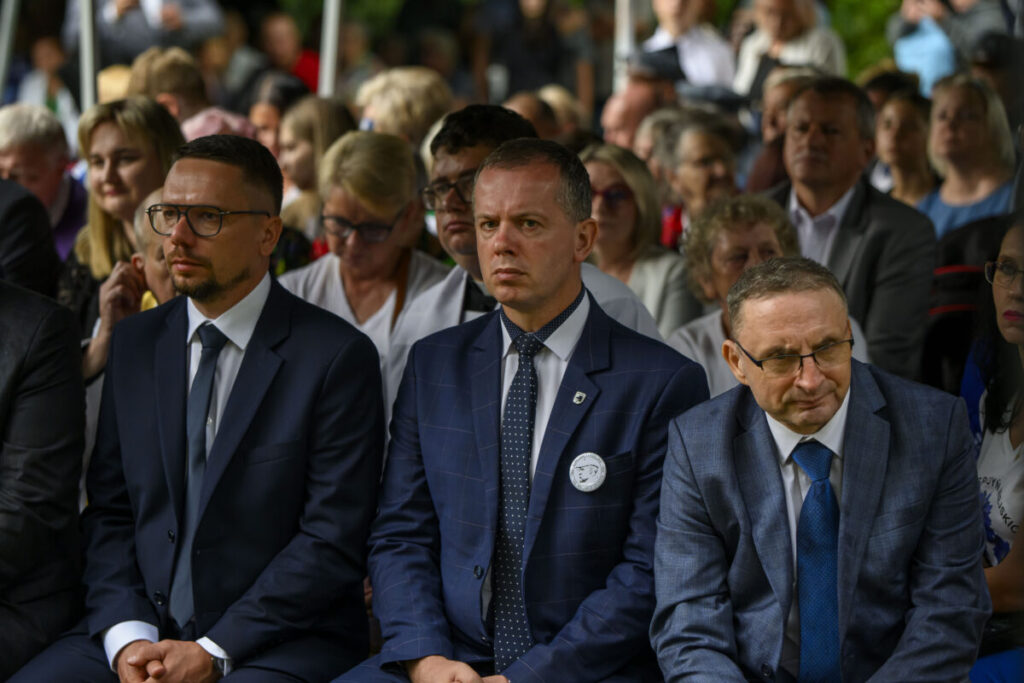 Grupa ludzi w garniturach siedzi w rzędzie na wydarzeniu. W tle widać innych uczestników. Atmosfera jest formalna.