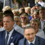 Grupa ludzi siedzi w rzędach; kobieta z różowymi okularami na środku. Tło jest rozmyte, dominuje kolor zielony i biały.