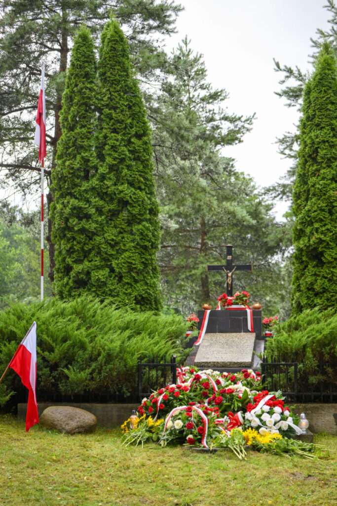 Grób z krzyżem na tle zieleni, otoczony wieńcami biało-czerwonych kwiatów. Flaga Polski po lewej. W tle drzewa.
