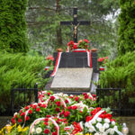 Grób z krzyżem otoczony wieńcami z czerwonych i białych kwiatów. Tło to zielone drzewa i krzewy. Kamień nagrobny z tablicą.