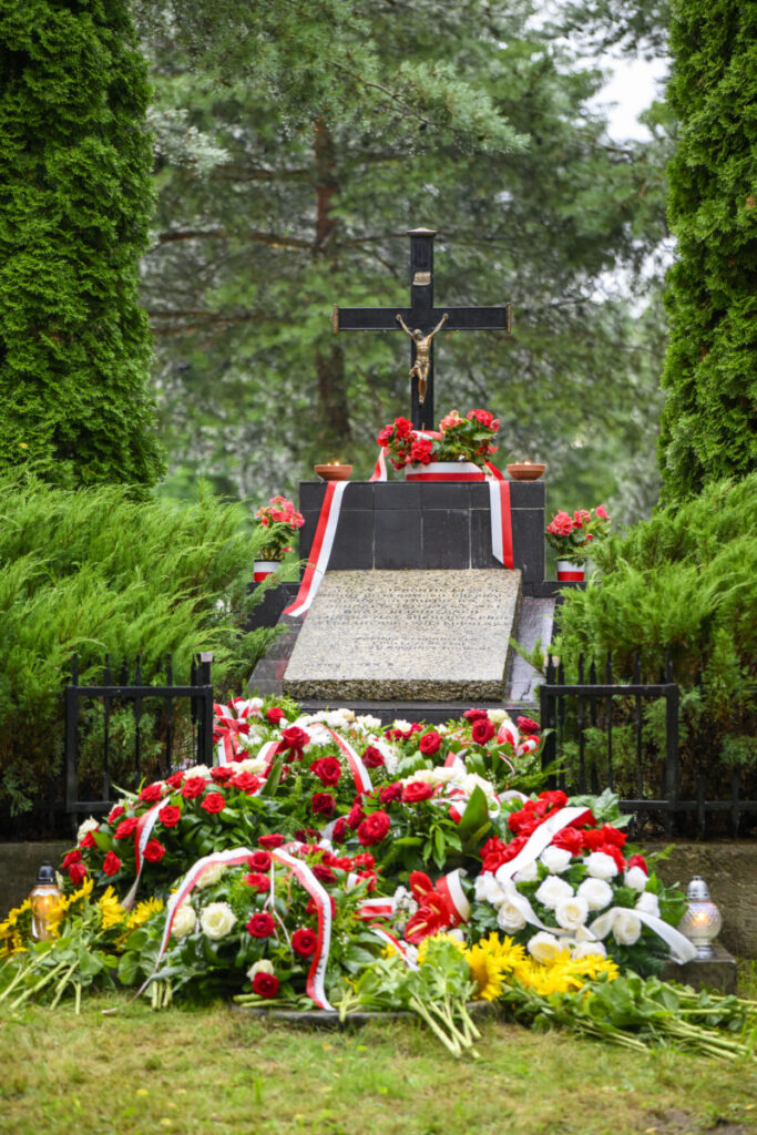 Grób z krzyżem otoczony wieńcami z czerwonych i białych kwiatów. Tło to zielone drzewa i krzewy. Kamień nagrobny z tablicą.