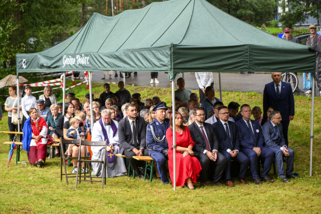 Na zdjęciu widoczna jest grupa ludzi pod zielonym namiotem na trawie. Osoby siedzą na ławkach, niektóre w formalnych strojach. Tło stanowi zieleń i asfaltowa droga.