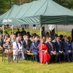 Na zdjęciu widoczna jest grupa ludzi pod zielonym namiotem na trawie. Osoby siedzą na ławkach, niektóre w formalnych strojach. Tło stanowi zieleń i asfaltowa droga.