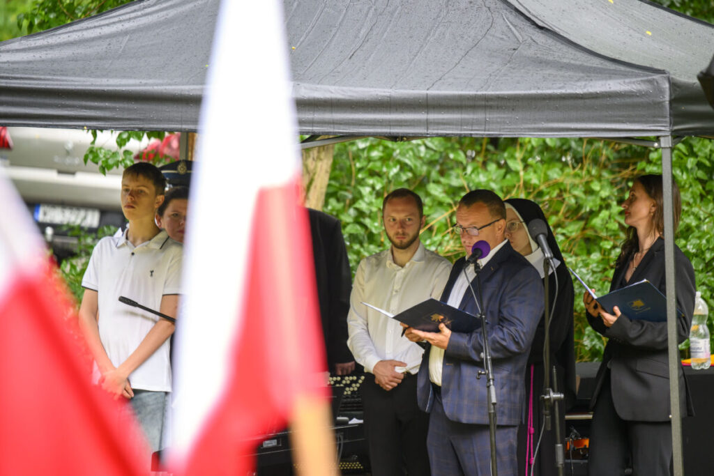 Grupa ludzi stoi pod czarnym namiotem. Mężczyzna przemawia do mikrofonu, trzymając książkę, otoczony osobami w eleganckich strojach. W tle zielone krzewy, na pierwszym planie polska flaga.