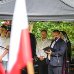 Grupa ludzi stoi pod czarnym namiotem. Mężczyzna przemawia do mikrofonu, trzymając książkę, otoczony osobami w eleganckich strojach. W tle zielone krzewy, na pierwszym planie polska flaga.