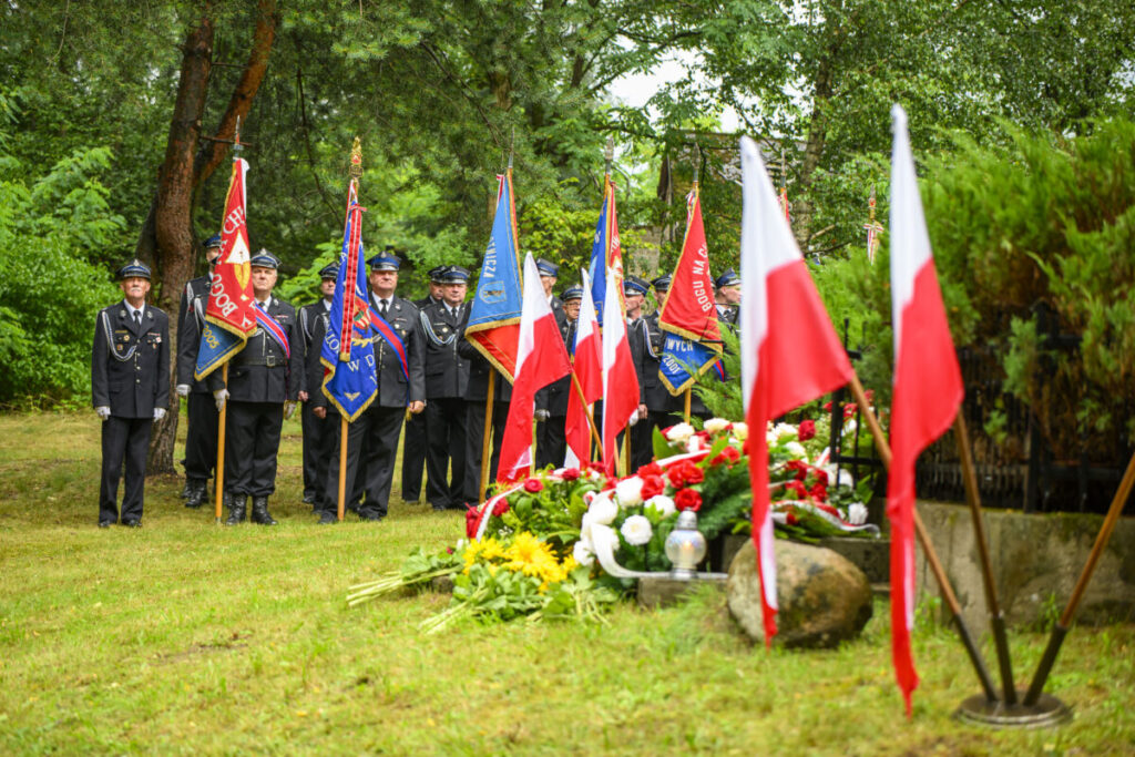 Ceremonia w lesie z uczestnikami w mundurach i flagami w kolorach Polski. W tle zielone drzewa. Na przodzie kwiaty i znicze.