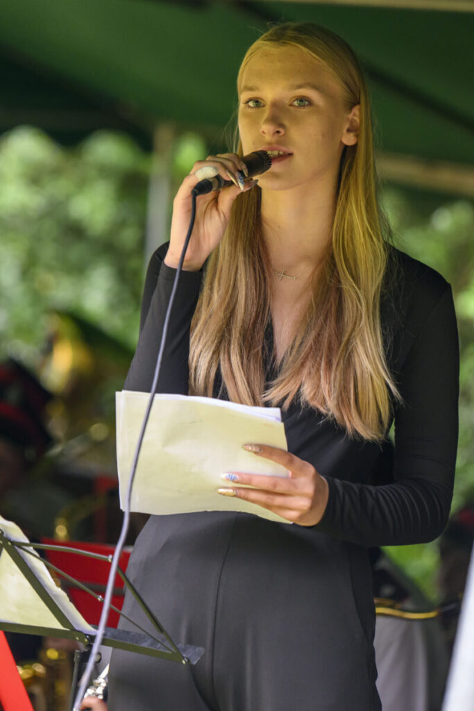 Osoba trzyma mikrofon i notatki, śpiewając. Ubrana na czarno, w tle rozmyta zieleń. Występ na świeżym powietrzu.