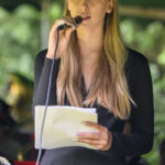 Nie wiem, kto jest na zdjęciu. Młoda kobieta w czarnej sukience śpiewa do mikrofonu, trzymając kartki. Tło zielone, rozmyte.