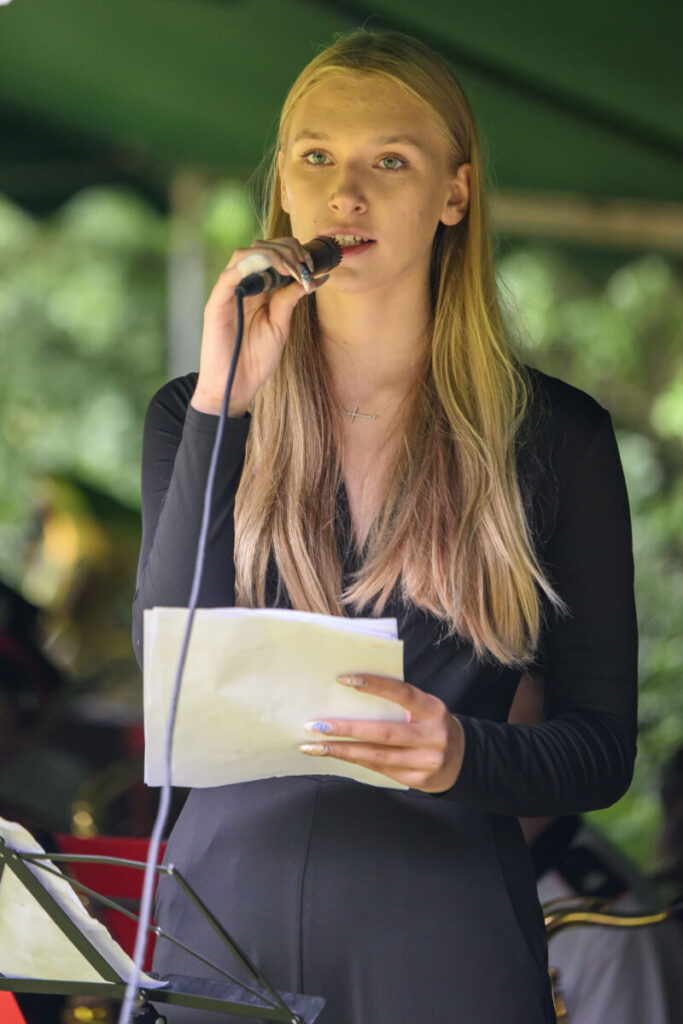 Nie wiem, kto jest na zdjęciu. Młoda kobieta w czarnej sukience śpiewa do mikrofonu, trzymając kartki. Tło zielone, rozmyte.