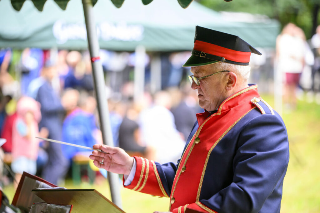 Mężczyzna w czerwono-granatowym mundurze dyryguje orkiestrą na świeżym powietrzu. Tło rozmyte, widoczni ludzie i zielony namiot. Sceneria festiwalowa.