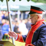 Mężczyzna w czerwono-granatowym mundurze dyryguje orkiestrą na świeżym powietrzu. Tło rozmyte, widoczni ludzie i zielony namiot. Sceneria festiwalowa.