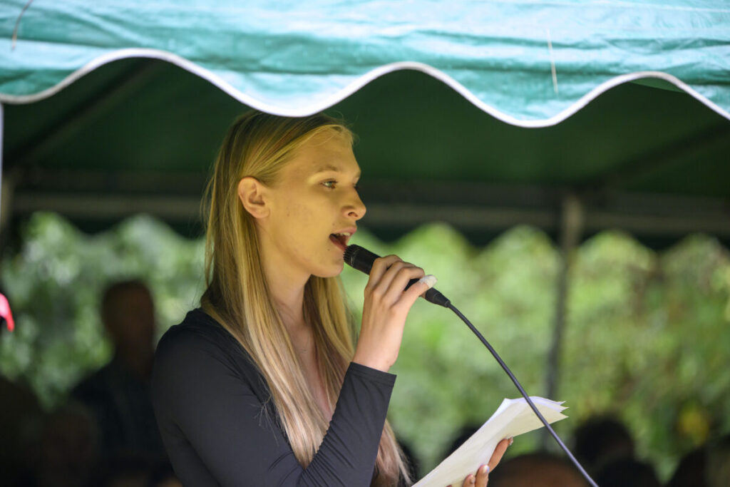 Młoda kobieta przemawia do mikrofonu, trzymając kartkę. Ma na sobie czarną bluzkę. W tle zielone zadaszenie i drzewa.