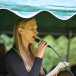 Młoda kobieta przemawia do mikrofonu, trzymając kartkę. Ma na sobie czarną bluzkę. W tle zielone zadaszenie i drzewa.