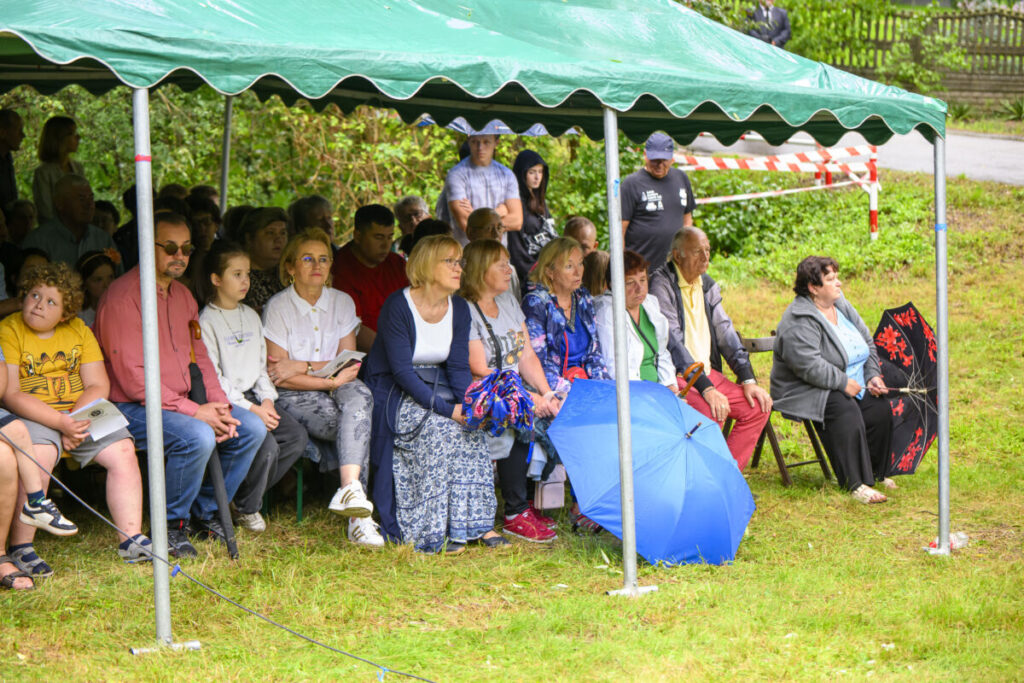 Grupa ludzi siedzi pod zielonym namiotem, na trawie, z otwartym niebieskim parasolem. W tle zielone liście i park. Atmosfera jest spokojna.