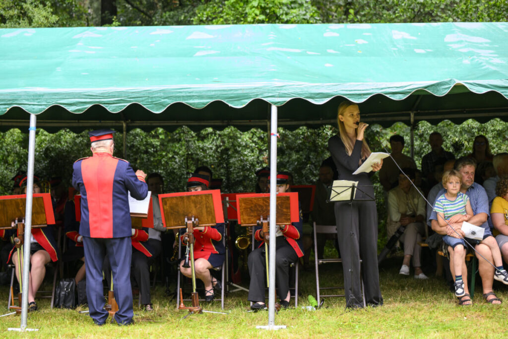 Zespół muzyczny w czerwonych mundurach gra pod zielonym namiotem. Dyrygent stoi tyłem, obok kobieta mówi do mikrofonu. Publiczność siedzi w tle.