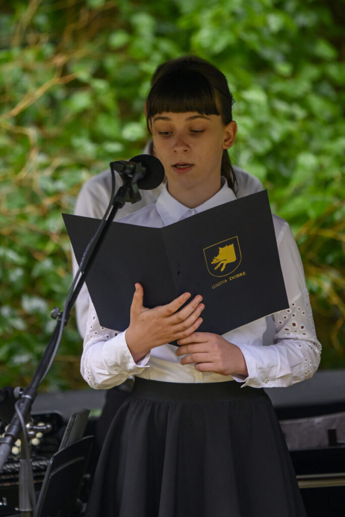 Na zdjęciu młoda osoba czyta z czarnej teczki, nosi białą koszulę. Tło stanowi zielone liście, a osoba stoi przy mikrofonie. Po prawej widać fragment instrumentu klawiszowego.