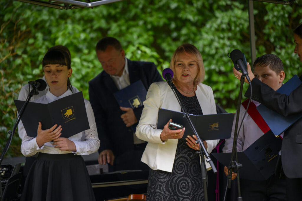 Grupa osób na scenie czyta z teczek. W tle zielone liście. Kolorystyka: biel, czerń, granat.