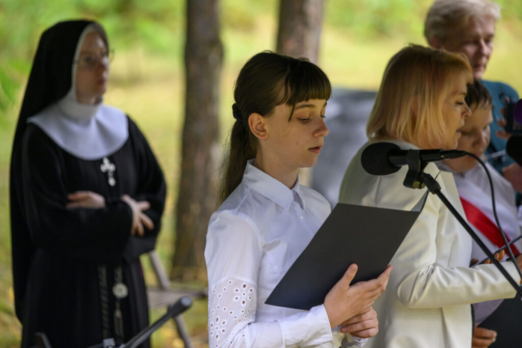Na zdjęciu młoda kobieta w białej koszuli z czarną teczką stoi przy mikrofonie. W tle widać zakonnicę w habicie i inne osoby. Tło jest zielone, z drzewami.