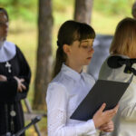 Na zdjęciu młoda kobieta w białej koszuli z czarną teczką stoi przy mikrofonie. W tle widać zakonnicę w habicie i inne osoby. Tło jest zielone, z drzewami.