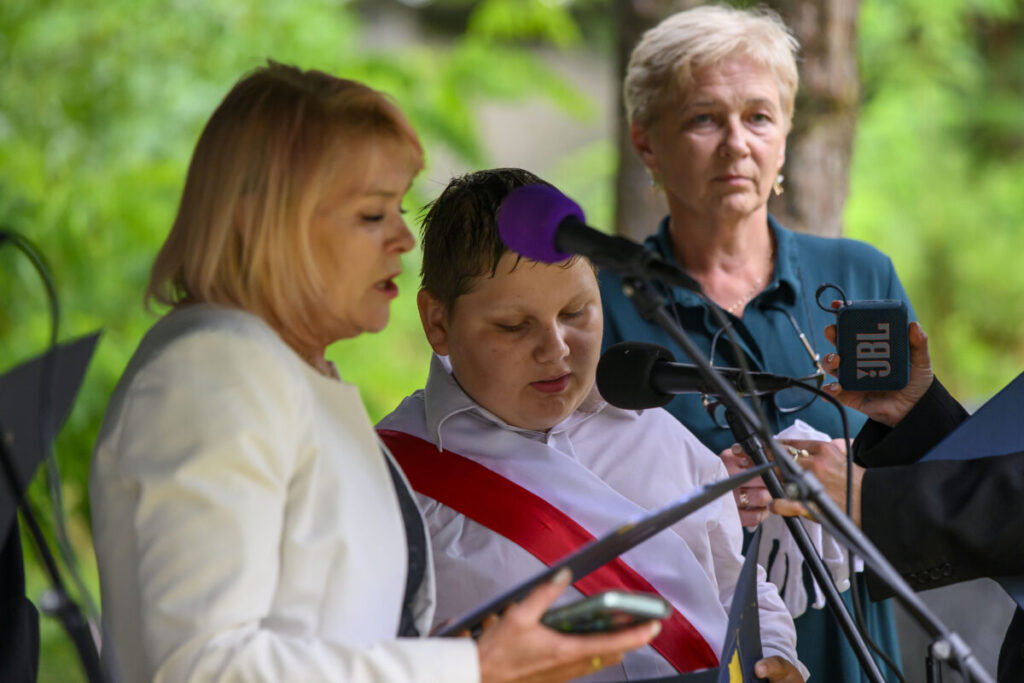 Na zdjęciu trzy osoby przy mikrofonie. Dziecko z białą koszulą i czerwoną szarfą mówi. Zielone tło drzew w tle.