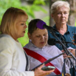 Na zdjęciu trzy osoby przy mikrofonie. Dziecko z białą koszulą i czerwoną szarfą mówi. Zielone tło drzew w tle.