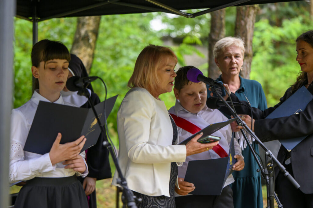 Grupa ludzi stoi pod zadaszeniem, trzymając notatki przy mikrofonach. W tle zielone drzewa. Kobieta w białej marynarce.
