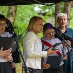 Grupa ludzi stoi pod zadaszeniem, trzymając notatki przy mikrofonach. W tle zielone drzewa. Kobieta w białej marynarce.