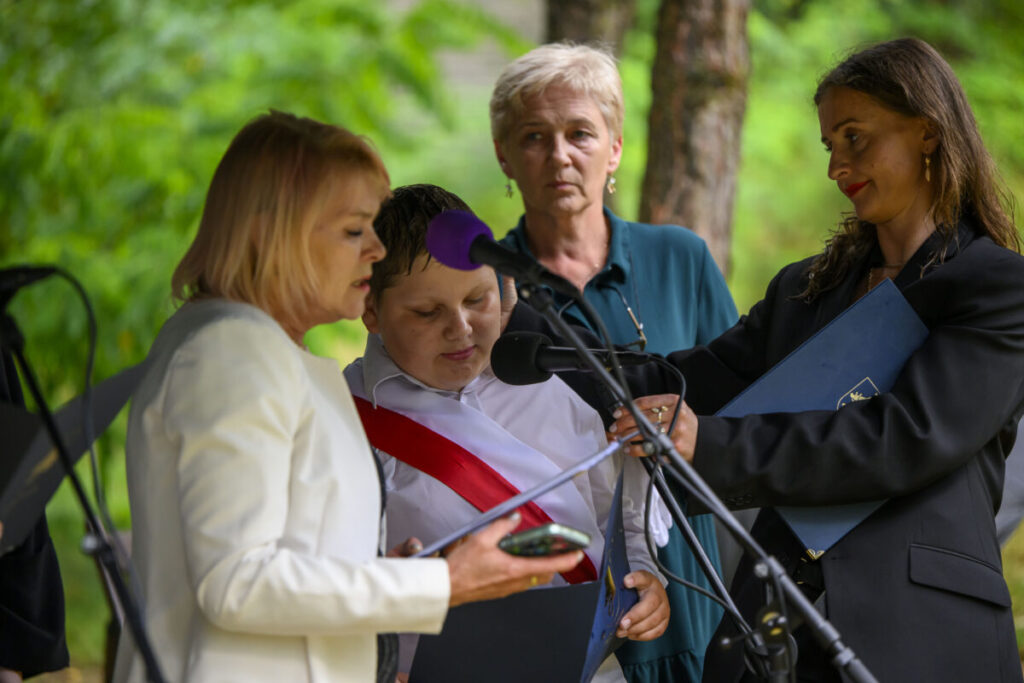 Na zdjęciu są cztery osoby przy mikrofonie na tle zieleni. Jedna osoba trzyma dokument, inna nosi białą koszulę z czerwoną wstęgą. Otoczenie jest naturalne, tło stanowi zieleń liści.