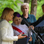 Na zdjęciu są cztery osoby przy mikrofonie na tle zieleni. Jedna osoba trzyma dokument, inna nosi białą koszulę z czerwoną wstęgą. Otoczenie jest naturalne, tło stanowi zieleń liści.