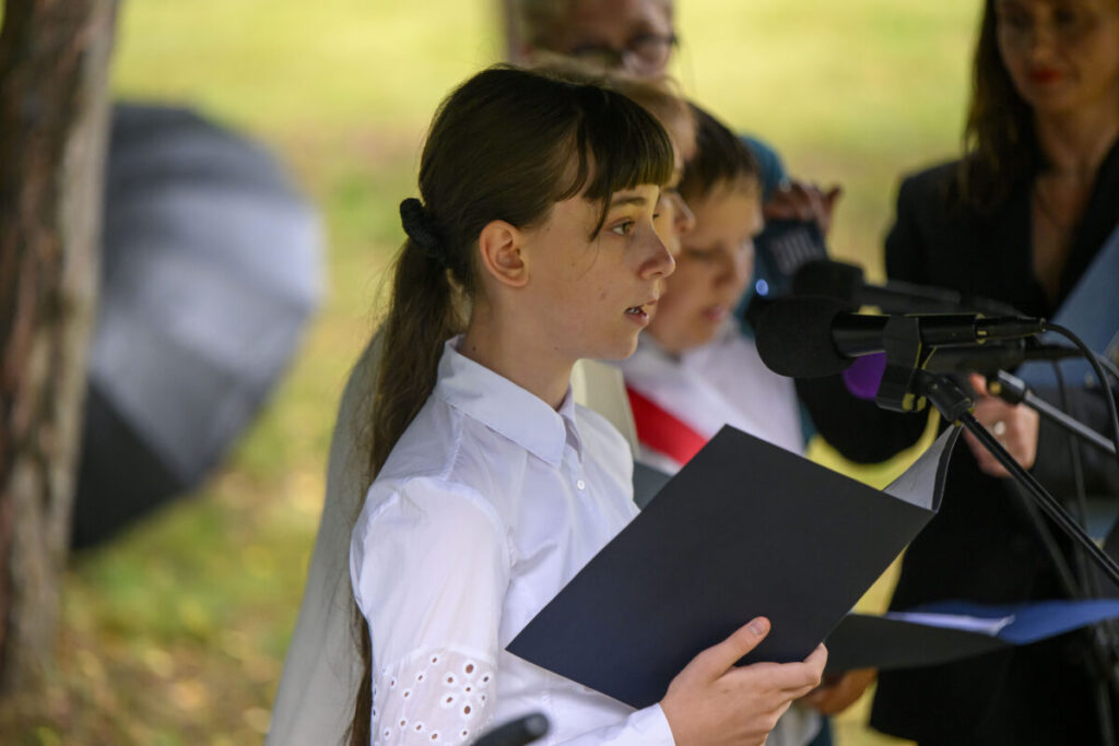 Na zdjęciu młoda osoba w białej koszuli czyta z kartki przy mikrofonie. Tło jest rozmazane, przypomina park. Widać parasol i kilka innych osób.
