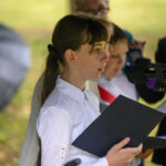 Na zdjęciu młoda osoba w białej koszuli czyta z kartki przy mikrofonie. Tło jest rozmazane, przypomina park. Widać parasol i kilka innych osób.
