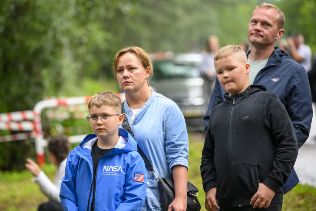 Grupa osób stoi na zewnątrz na tle zieleni. Przeważają niebieskie i czarne ubrania. Tło jest rozmyte, a ogrodzenie w tle ma biało-czerwone taśmy.