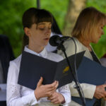 Osoba w białej koszuli czyta z czarnej teczki przy mikrofonie. W tle zieleń drzew. Obok stoi inna osoba.