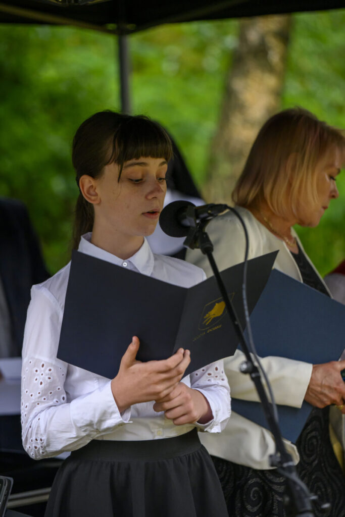 Osoba w białej koszuli czyta z czarnej teczki przy mikrofonie. W tle zieleń drzew. Obok stoi inna osoba.