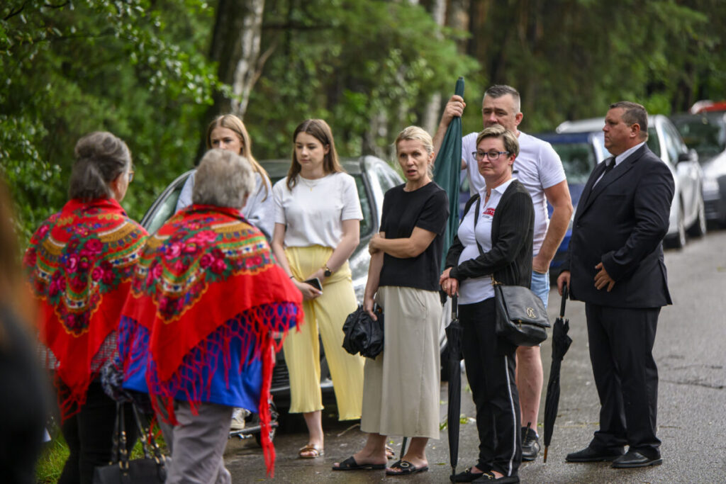 Grupa ludzi stoi na ulicy, w tle drzewa. Dwie osoby w czerwonych chustach z kwiatami. Kilka osób trzyma parasole.