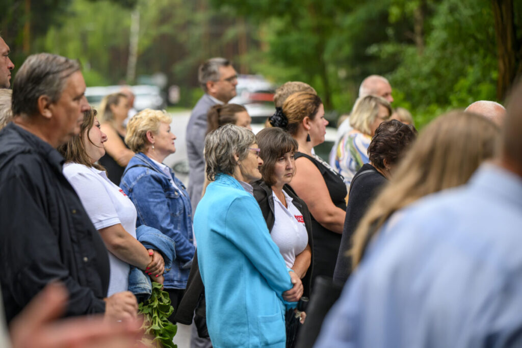 Ludzie stoją w skupieniu na zewnątrz, w otoczeniu zieleni. Przeważają ubrania w kolorach niebieskim i białym. Tło stanowi zamazana zieleń drzew.