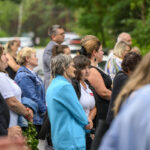 Ludzie stoją w skupieniu na zewnątrz, w otoczeniu zieleni. Przeważają ubrania w kolorach niebieskim i białym. Tło stanowi zamazana zieleń drzew.