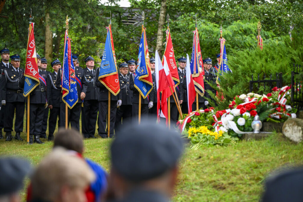 Grupa strażaków w mundurach stoi z kolorowymi sztandarami na tle zielonych drzew. Na pierwszym planie widoczne są biało-czerwone kwiaty. Tło jest zielone, z drzewami i trawą.