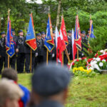 Grupa strażaków w mundurach stoi z kolorowymi sztandarami na tle zielonych drzew. Na pierwszym planie widoczne są biało-czerwone kwiaty. Tło jest zielone, z drzewami i trawą.