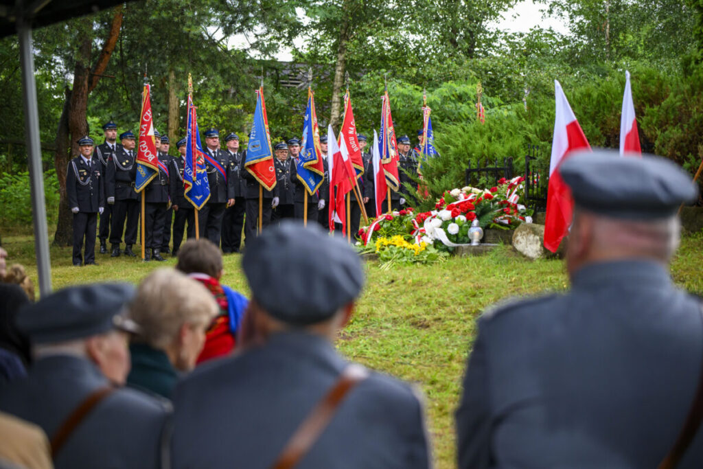 Uroczystość z udziałem osób w mundurach i wieloma flagami. Czerwone i białe kwiaty leżą obok pomnika. W tle zielone drzewa.