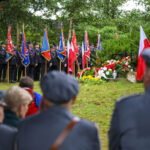 Uroczystość z udziałem osób w mundurach i wieloma flagami. Czerwone i białe kwiaty leżą obok pomnika. W tle zielone drzewa.