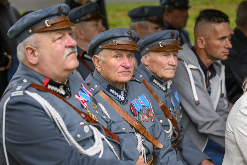 Grupa starszych mężczyzn w szarych mundurach z medalami siedzi w rzędzie. Mundury ozdobione są kolorowymi odznaczeniami. W tle rozmyte otoczenie natury.